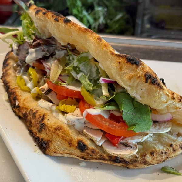 A Mediterranean flatbread sandwich overflowing with fresh vegetables, meat, and white sauce on a white plate.