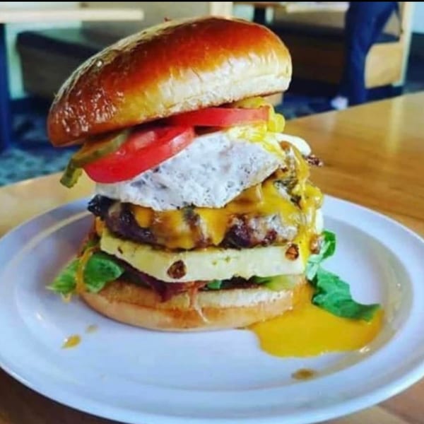 A tall burger with cheese, egg, pineapple, tomato, and pickles on a white plate.