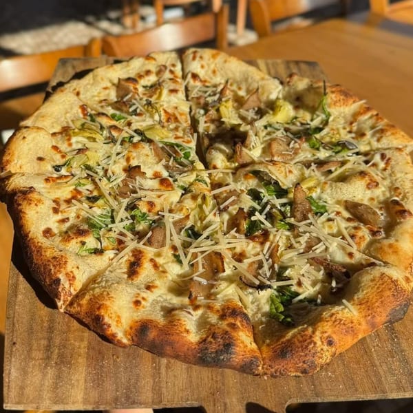 A Mediterranean pizza with white sauce, artichokes, sausage, and shredded cheese on a wooden board.