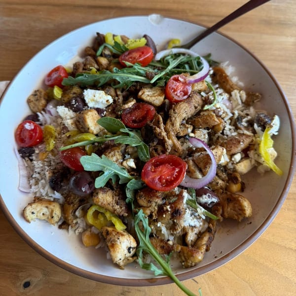 A Mediterranean bowl with chicken, rice, tomatoes, onions, feta, and arugula on a wooden table.